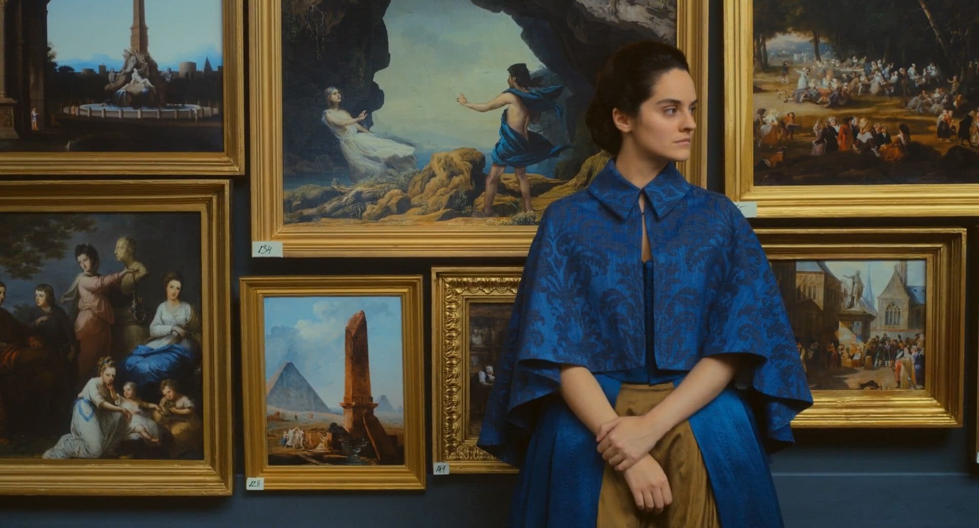Still from Portrait of a Lady on Fire (2019). Marianne, dressed in an elegant blue dress with a cropped cloak over her shoulders, stands in front of an art gallery wall. There are several gold-framed paintings behind her, the largest depicting the myth of Orpheus and Eurydice.