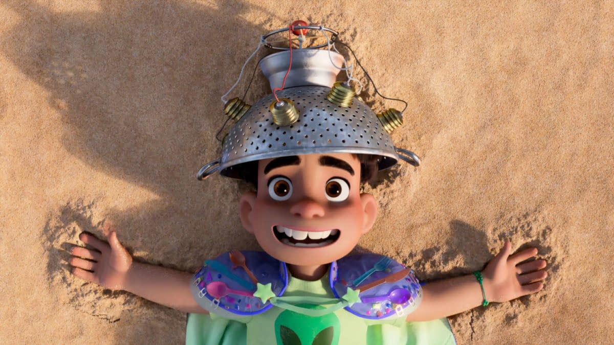 A scene from the animated film 'Elio' featuring a young boy with a cheerful expression lying on sandy ground, wearing a colander as a hat and colorful armor, embracing his imaginative and adventurous spirit. The film, produced by Disney and Pixar, explores themes of space and self-discovery.