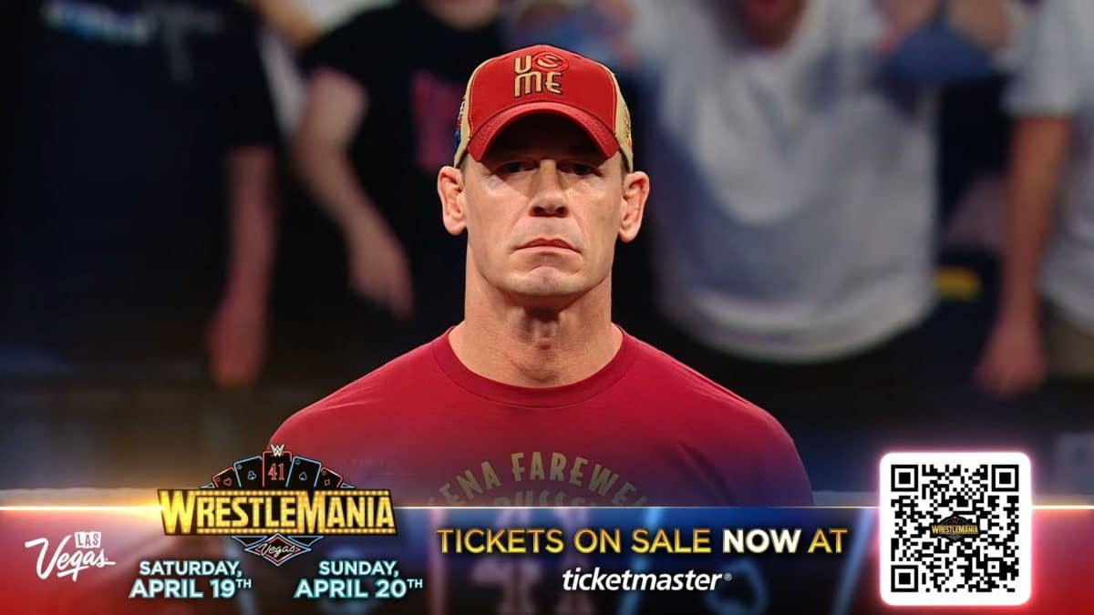 John Cena stands in the ring during WWE Raw in Brussels, wearing a red shirt and a red cap. The background shows blurred fans cheering.