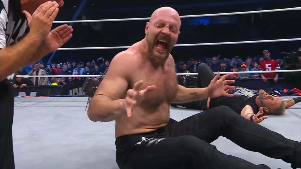 Jon Moxley, visibly in pain, sits in the wrestling ring with spikes protruding from his back after a confrontation with Adam Copeland. The referee is present, appearing to call for assistance.