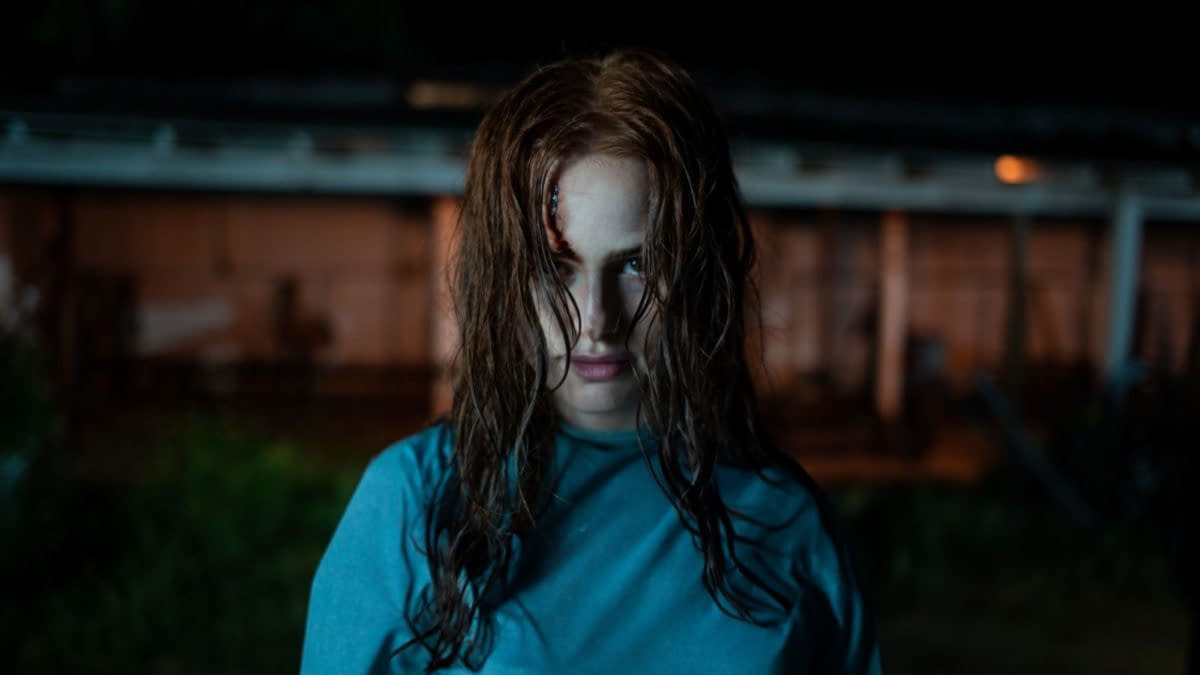 A woman with disheveled hair and an intense expression stands in a dimly lit outdoor setting, wearing a blue shirt. A small wound is visible on her forehead, adding to the eerie atmosphere.