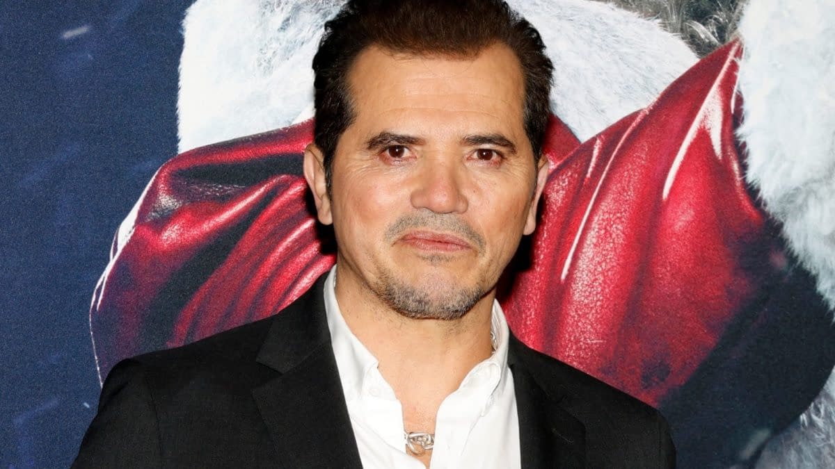 John Leguizamo poses on the red carpet at a movie premiere event in Hollywood, with a festive background featuring a Santa Claus image. He is dressed in a black suit and white shirt.