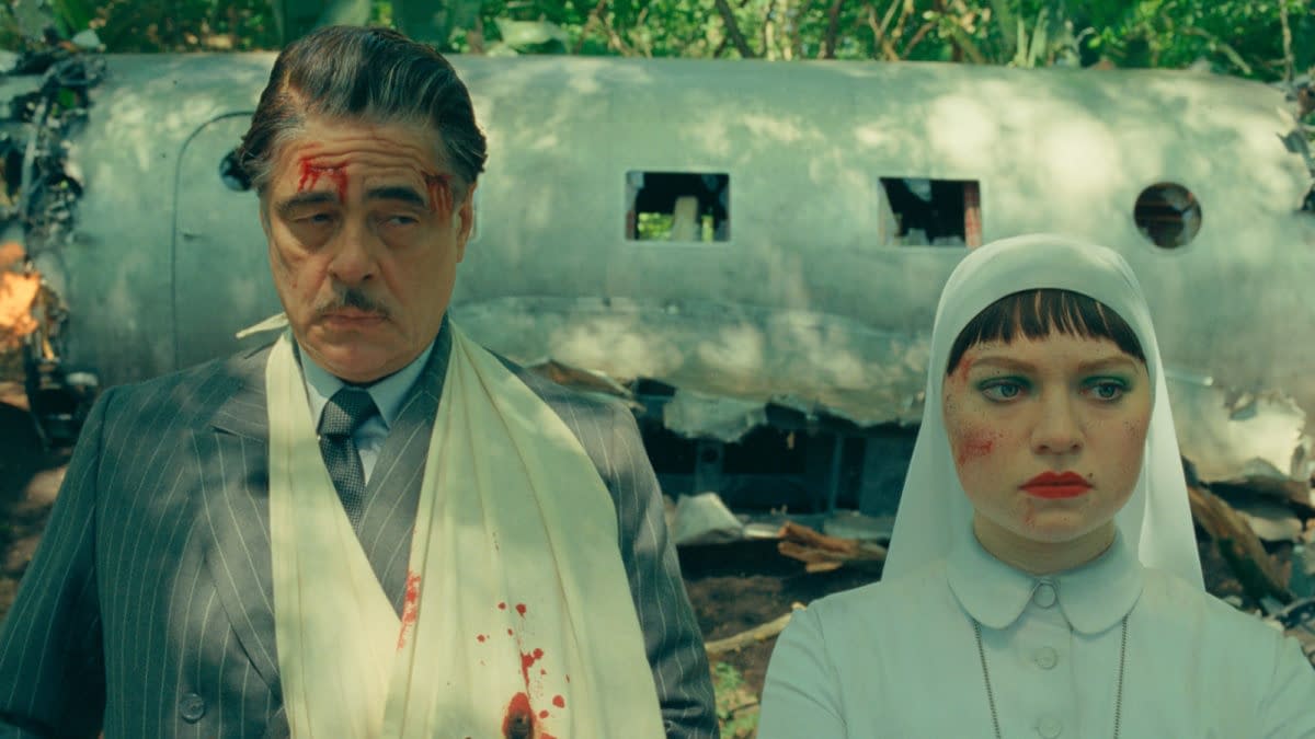 A still from the film "The Phoenician Scheme" featuring Benicio Del Toro as Zsa-Zsa Korda, wearing a blood-stained bandage, alongside Mia Threapleton as Liesl, who is dressed in a white outfit and a headscarf. The background shows a damaged airplane amid lush greenery.