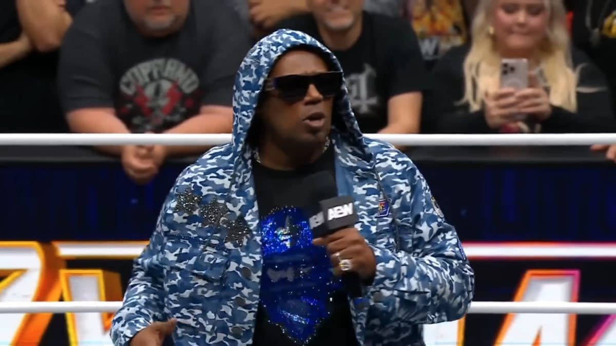 Master P, wearing a blue camo hoodie and sunglasses, is speaking into a microphone in a wrestling ring, engaging with the audience. Fans can be seen in the background, showing their excitement.