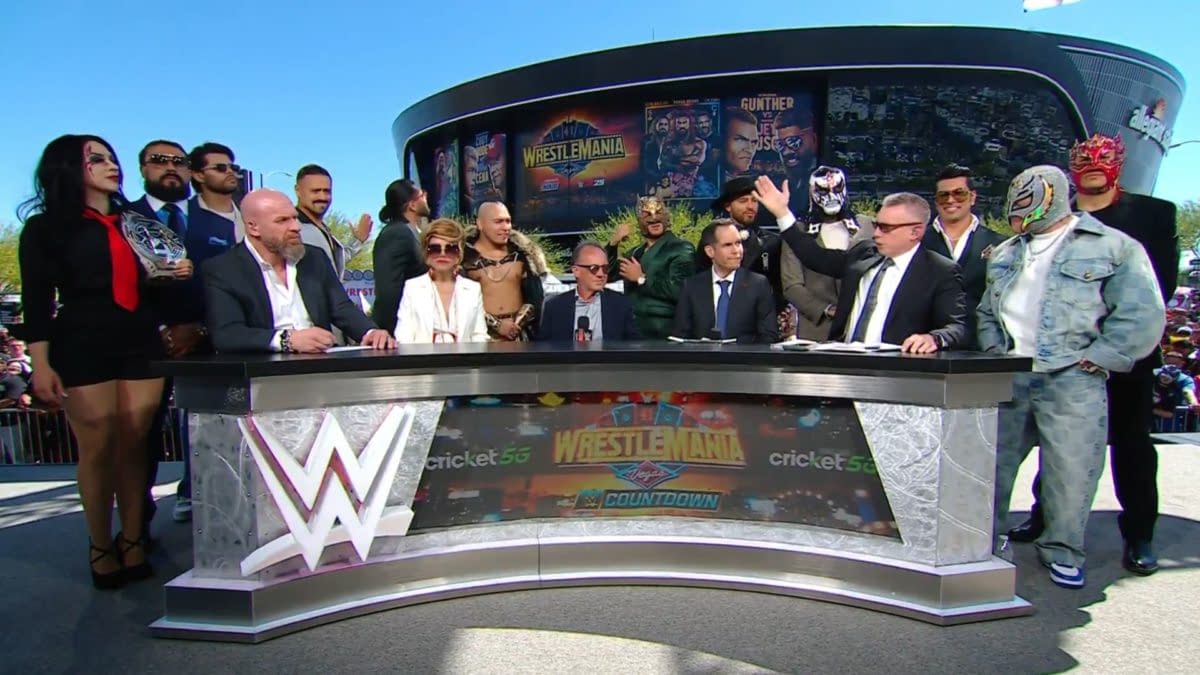 A group of wrestlers and executives from WWE and AAA stand behind a table during a press conference. They are gathered outdoors at WrestleMania 41 weekend in Las Vegas, showcasing diverse costumes and expressions of excitement.