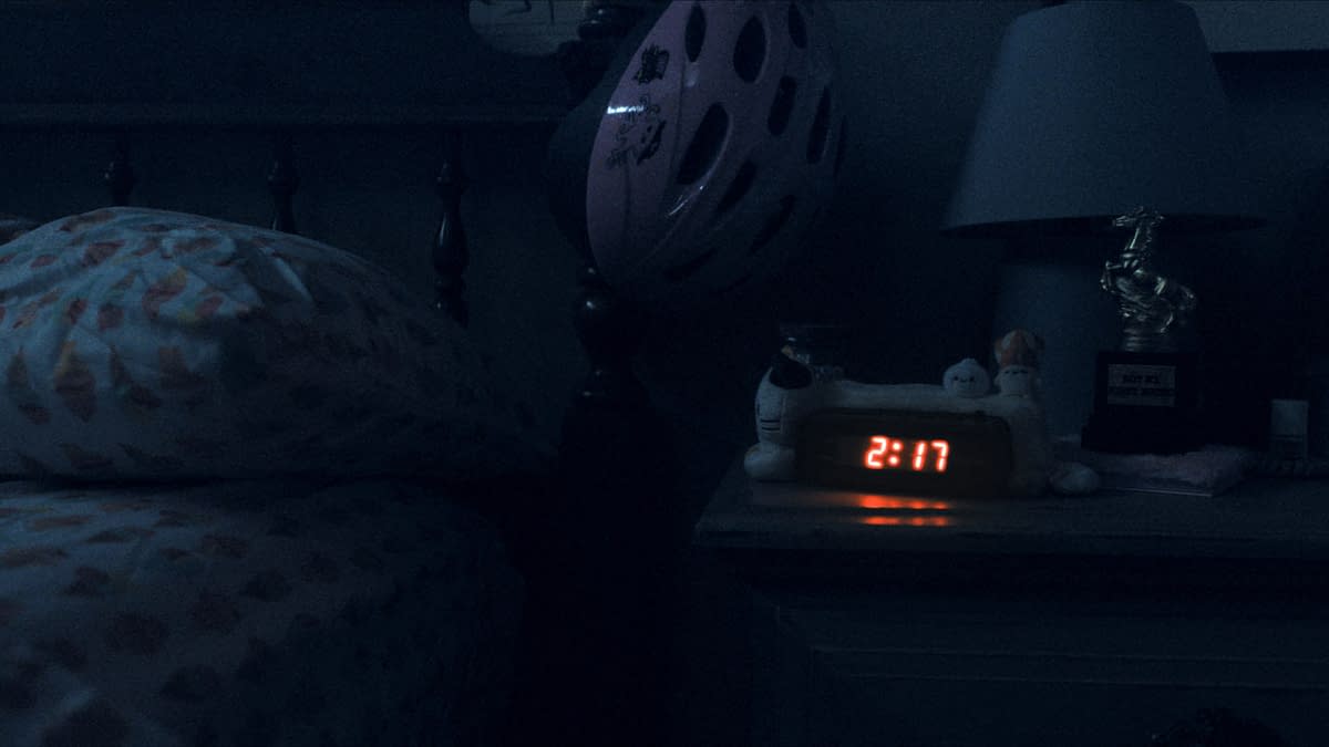A dimly lit bedroom at night featuring a bedside table with a digital clock displaying 2:17, various small plush toys, a lamp with a horse figurine, and a pink bicycle helmet hanging nearby.