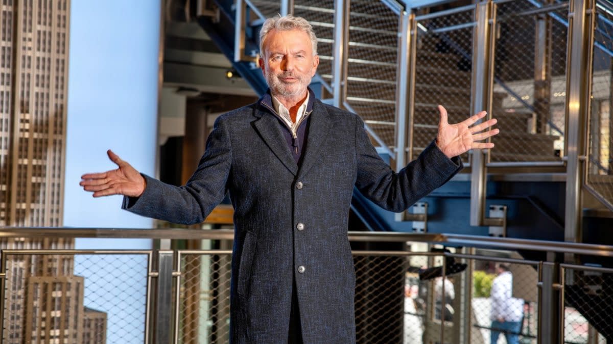Actor Sam Neill stands in the Empire State Building with arms open, showcasing the iconic architecture around him.