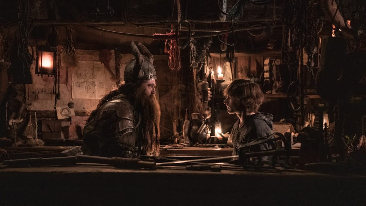 A scene from 'How To Train Your Dragon' featuring a bearded character in armor speaking to a young boy in a dimly lit workshop filled with various tools and decorations.