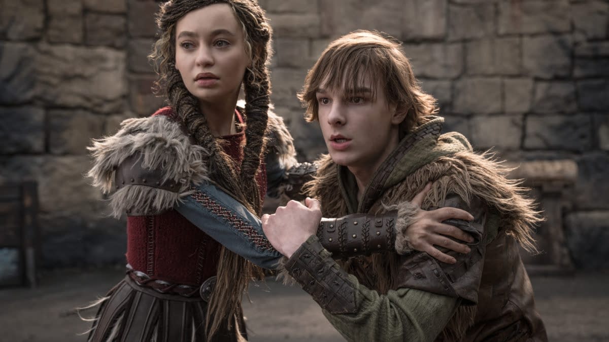 A scene from 'How To Train Your Dragon' featuring two characters, a young woman with braided hair in a red and grey costume, and a young man with tousled hair in a green hoodie and fur-trimmed clothing, looking distressed while holding onto each other in a medieval setting.