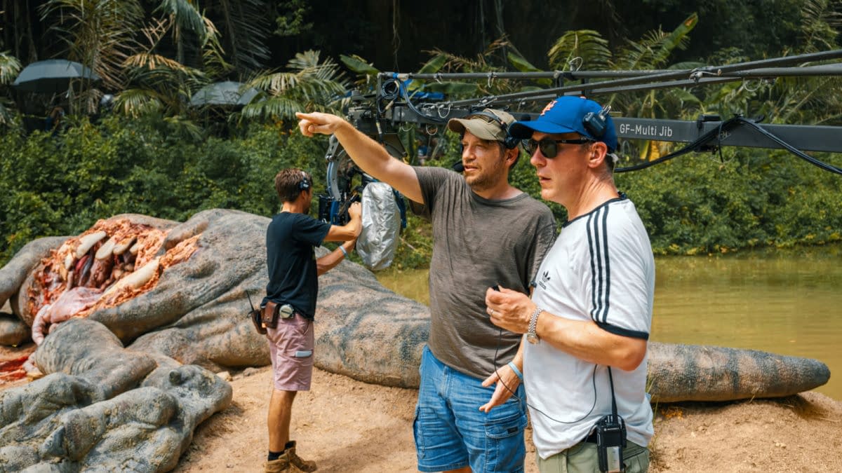 On the set of 'Jurassic World Rebirth,' director Gareth Edwards points while discussing a scene, flanked by crew members and a large, dinosaur-like prop in a lush, green environment.