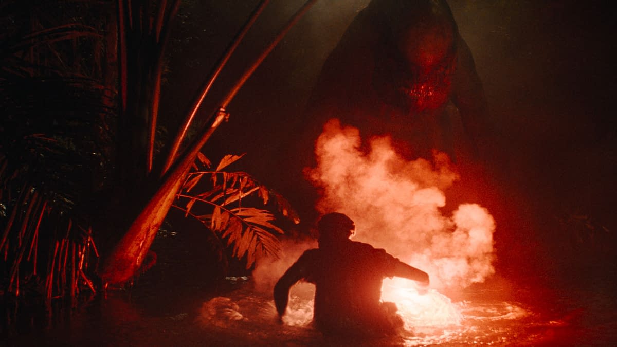 A tense scene from 'Jurassic World Rebirth,' featuring a silhouetted figure in water, illuminated by a glowing object, with a menacing dinosaur emerging in the background, surrounded by dark foliage and smoke.
