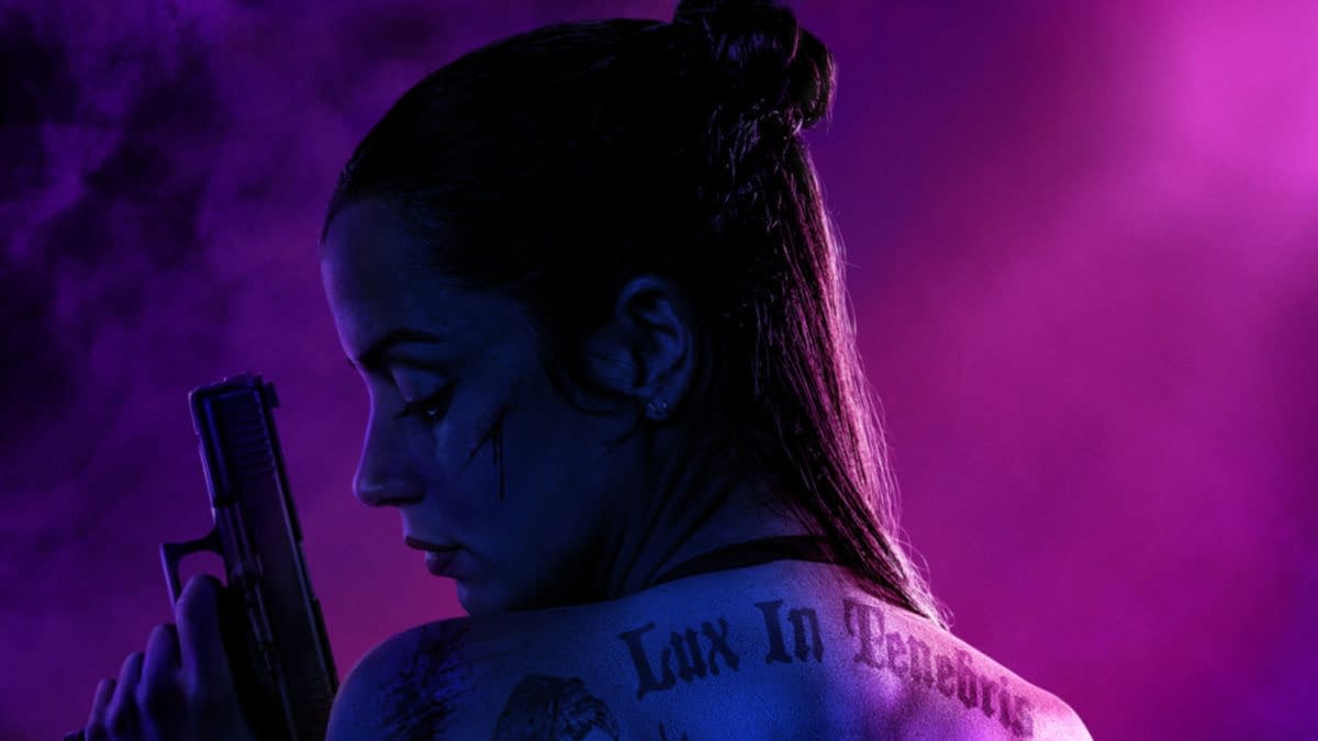 A woman with long, dark hair styled in a bun poses with a gun, her back to the viewer. Her tattooed shoulder features the phrase 'Lux In Tenebris,' while dramatic purple lighting creates a moody atmosphere.