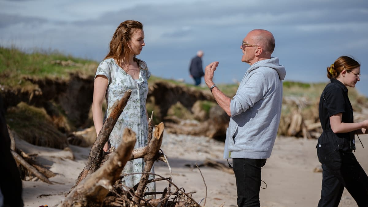 A behind-the-scenes moment on a seaside set, featuring a woman in a floral dress engaging in conversation with a man wearing a gray hoodie. A crew member is seen in the background, alongside driftwood scattered on the beach.