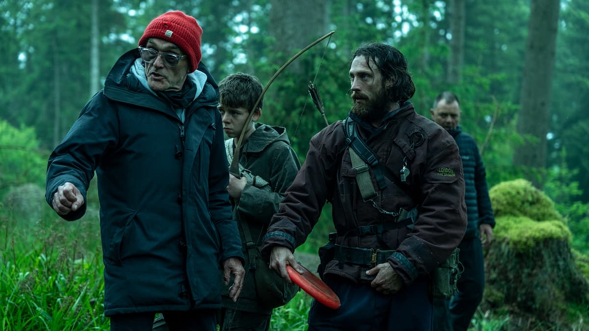 A group of four people in a forest, with one wearing a red beanie and gesturing, while another holds an arrow and bow. The scene conveys a sense of urgency and focus, suitable for a narrative about survival or adventure.
