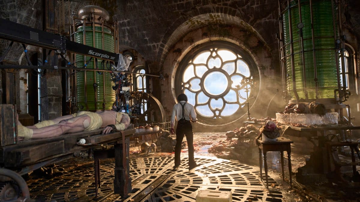 On the set of Frankenstein, Jacob Elordi portrays The Creature while Oscar Isaac as Dr. Victor Frankenstein stands in a dimly lit laboratory with a large stained glass window. The scene features a dramatic atmosphere with scientific equipment and red lighting, emphasizing themes of creation and destruction.