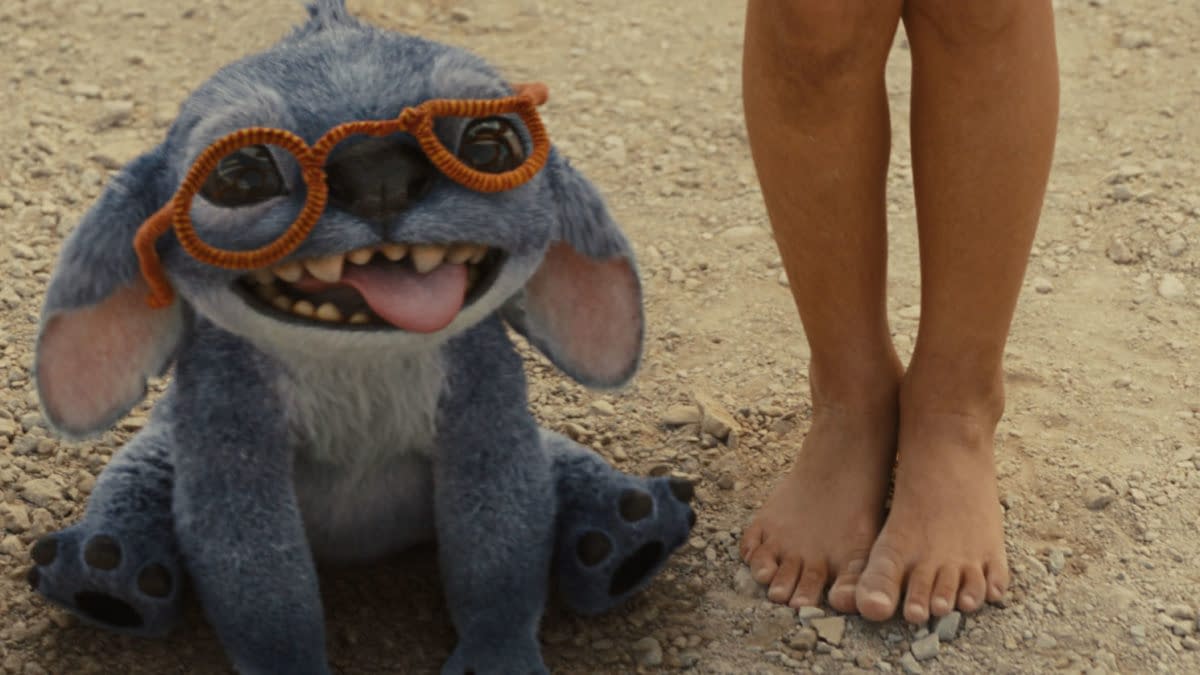 Stitch, a blue alien character from Disney's live-action 'Lilo & Stitch,' is sitting on the ground, wearing orange glasses and playfully sticking out his tongue alongside a child's bare feet.