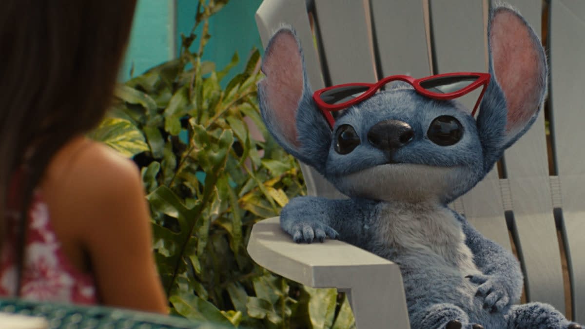 In Disney's live-action Lilo & Stitch, Stitch, the blue alien character, wears red sunglasses and relaxes in a white lawn chair, facing Lilo, who is partially visible in the background surrounded by greenery.
