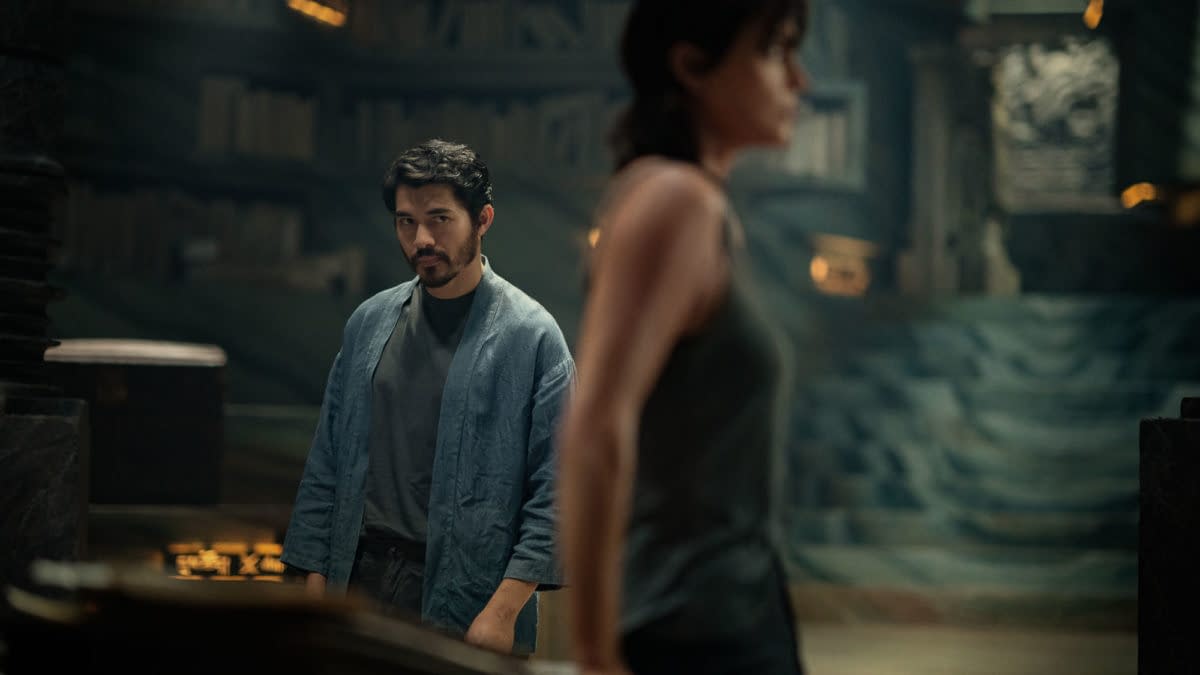 A scene from 'The Old Guard 2' featuring Henry Golding as Tuah in a blue robe, looking intently at Charlize Theron as Andy, who is in the foreground. The setting is dimly lit with shelves of books in the background.