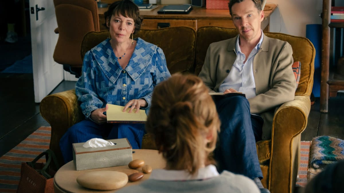 A scene from 'The Roses' featuring Olivia Colman and Benedict Cumberbatch sitting on a couch, engaged in conversation with a person out of frame. The setting includes a cozy living room with a coffee table displaying a tissue box and smooth stones.