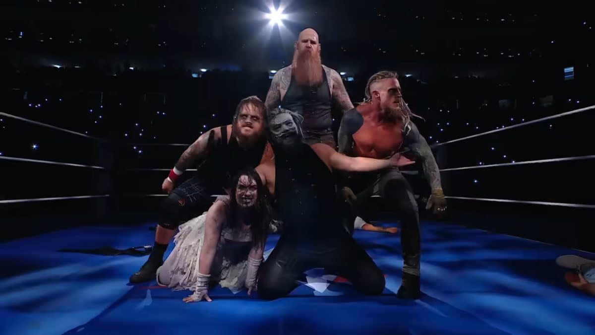 A group of wrestlers, dressed in spooky and dark attire, poses dramatically in a wrestling ring under bright lights, with a dark audience in the background.
