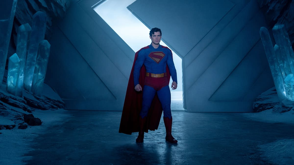 Superman stands confidently in a futuristic ice cave, wearing his iconic blue suit with a red cape. Crystalline structures are visible in the background against a blue-lit atmosphere.