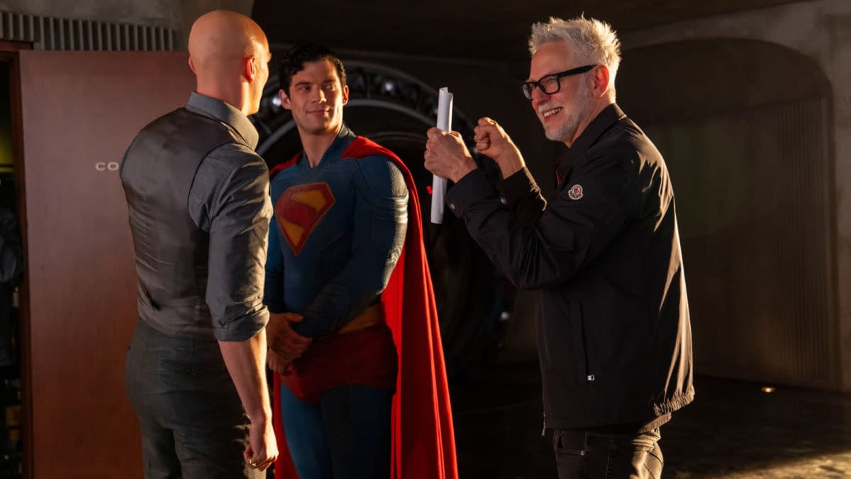 A behind-the-scenes moment featuring two men conversing, one dressed as Superman in a blue suit with a red cape, and a third man smiling and holding a script, likely a director on set.