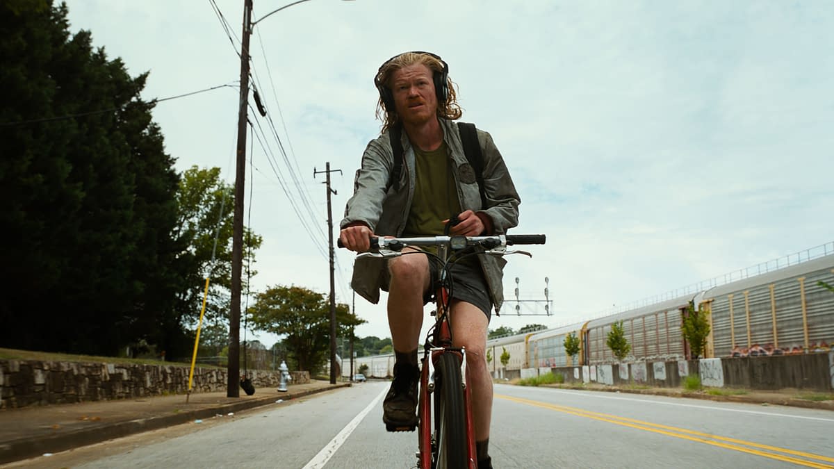 A scene from the film 'BUGONIA' (2025) featuring a man with long hair wearing headphones, riding a bicycle along a city street. He appears focused and is dressed in casual attire with a backpack.