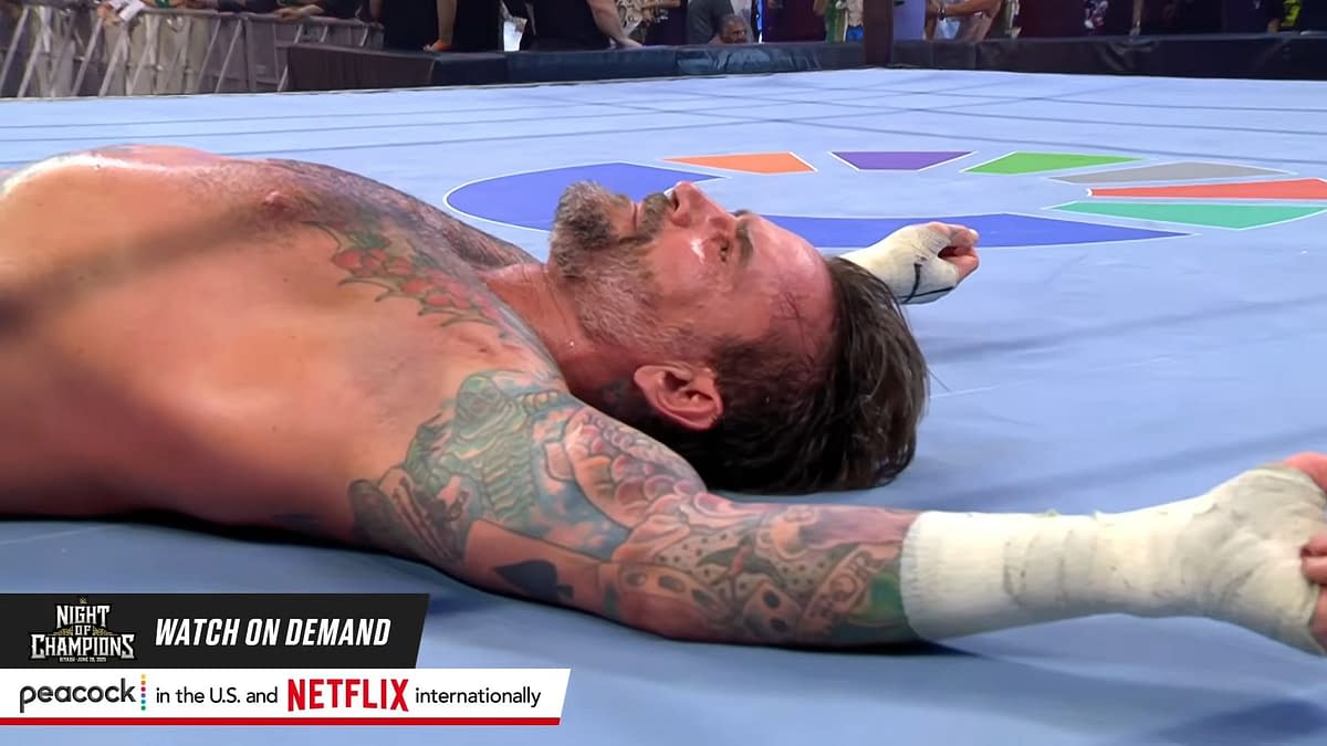 A defeated wrestler lies on the wrestling ring floor, showcasing tattoos on his arms and a weary expression. It is CM Punk in the aftermath of his match with John Cena at WWE Night of Champions in Saudi Arabia.