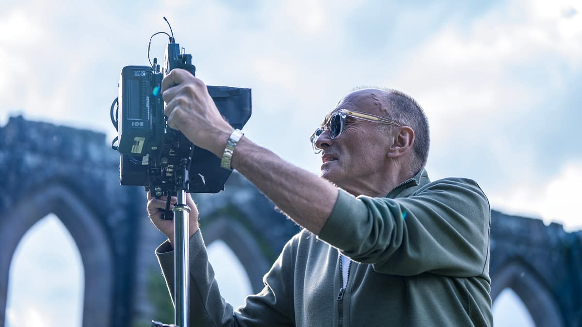A photographer, wearing sunglasses, is adjusting a camera on a tripod in front of a historic stone archway under a bright sky.