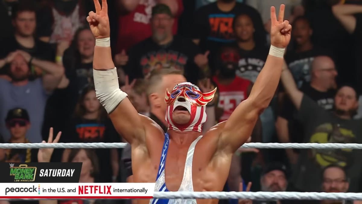 El Grande Americano, wearing a colorful mask and outfit, celebrates a victory in the wrestling ring, raising his arms in triumph. The excited crowd is visible in the background, reacting to the moment.