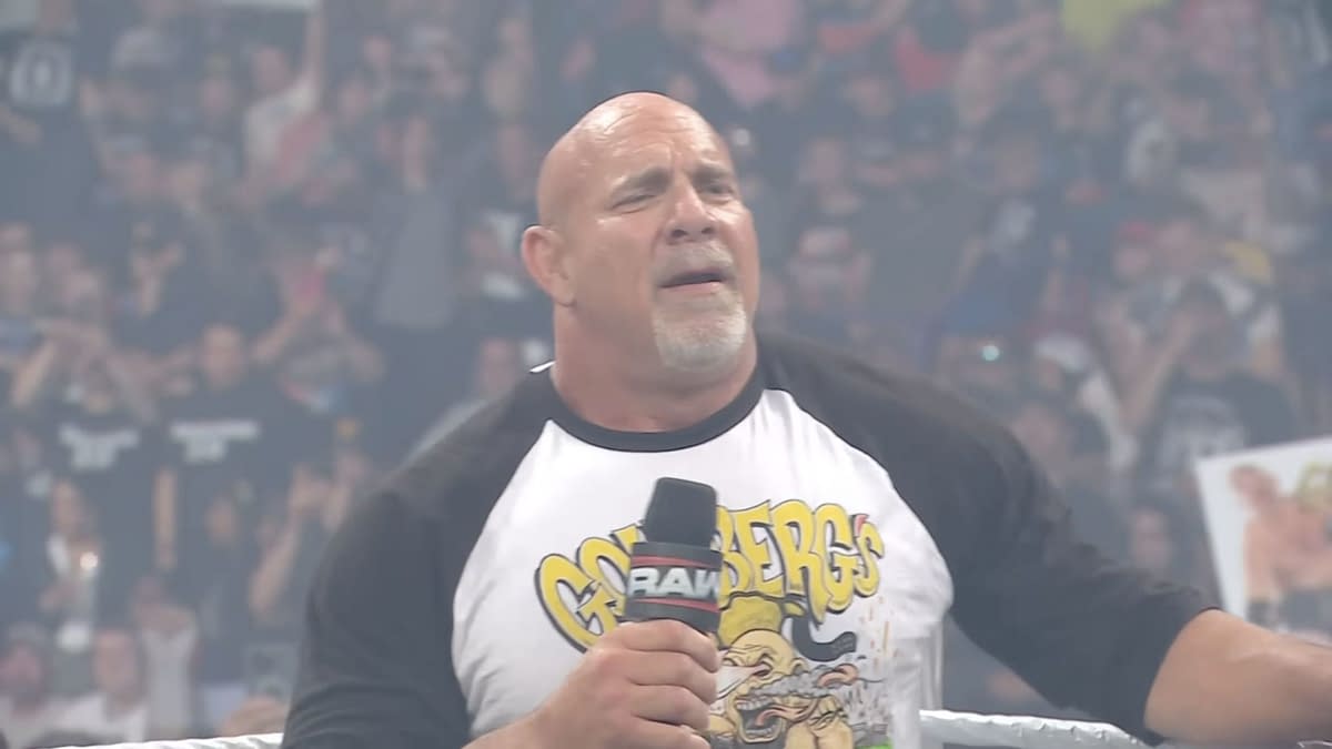 A muscular man in a 'Goldberg' t-shirt stands in a wrestling ring, holding a microphone. The crowd is visible in the background, cheering enthusiastically.