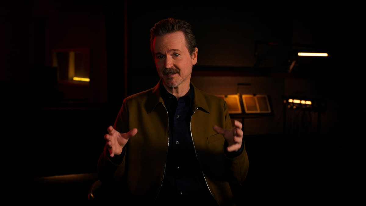 A man with a mustache speaks animatedly, wearing a brown jacket over a dark shirt, in a dimly lit setting with warm lighting. The background features a blurred view of books and instruments.