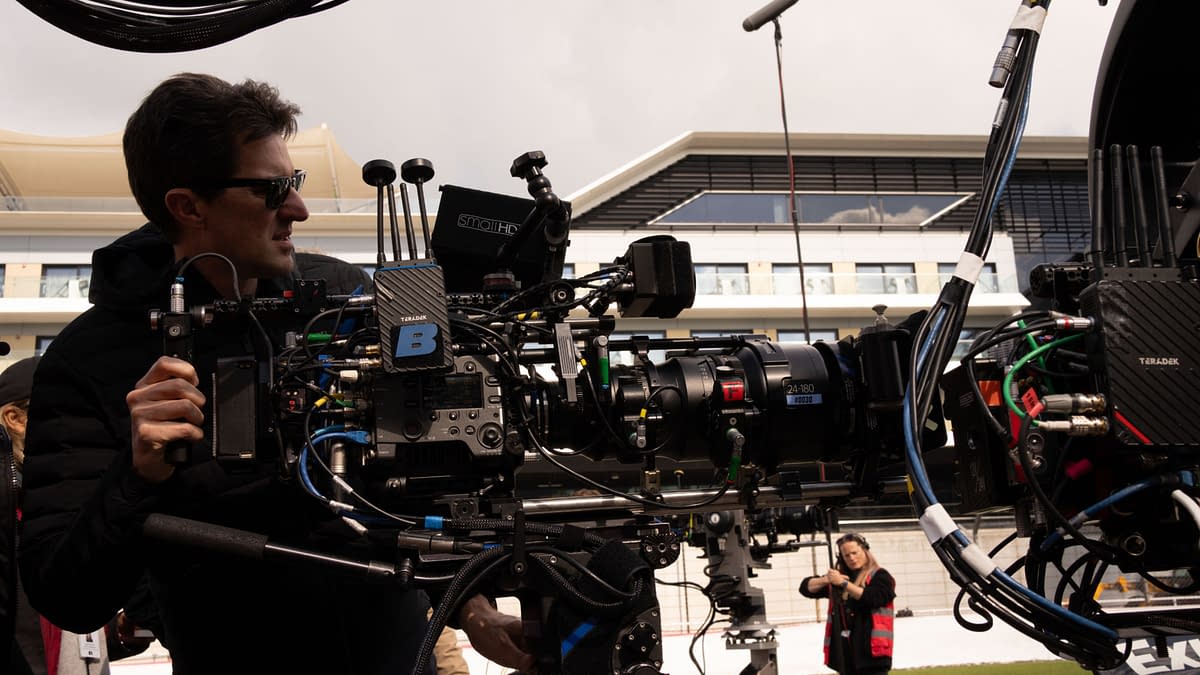 A film crew member operates a professional camera setup on a film set, with various cables and equipment visible, while another crew member prepares in the background. The setting appears to be an outdoor venue, suggesting a dynamic production environment.