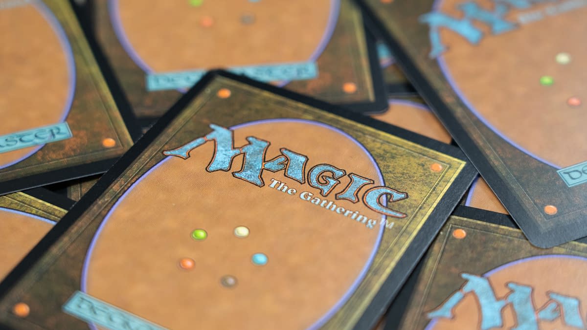 Colorful backs of Magic: The Gathering trading cards are stacked together, featuring an ornate design with a prominent logo and various colored dots, emphasizing the fantasy and gaming theme.