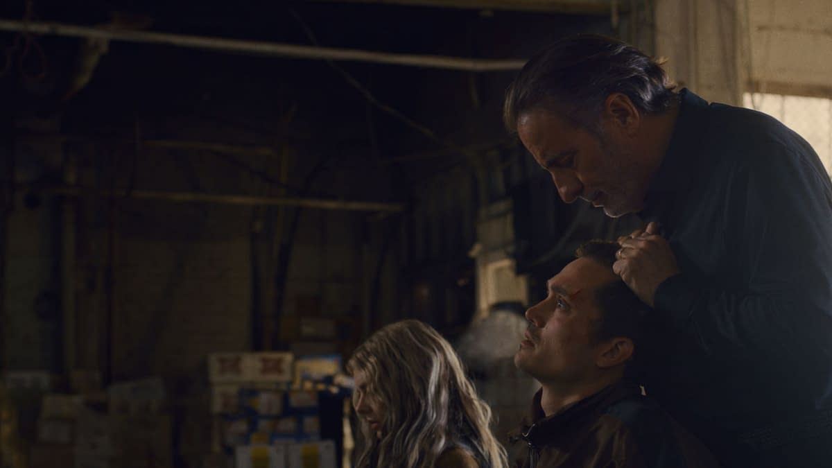 A tense scene from the film 'Eenie Meanie' featuring Samara Weaving as Edie, Karl Glusman as John, and Andy Garcia as Nico in a dimly lit industrial setting. The characters display intense emotions, with Nico holding John's head, suggesting a confrontation.