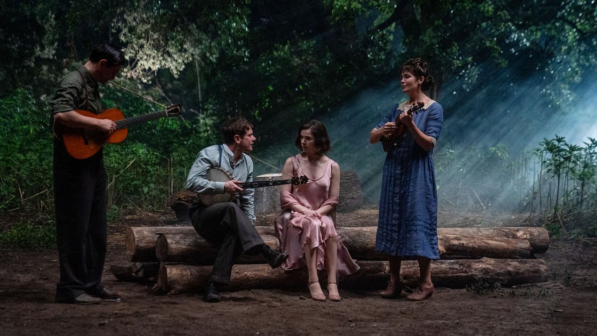 A scene featuring four performers in a forest setting, with two holding string instruments and two seated, creating a cozy atmosphere illuminated by soft, natural light filtering through the trees.