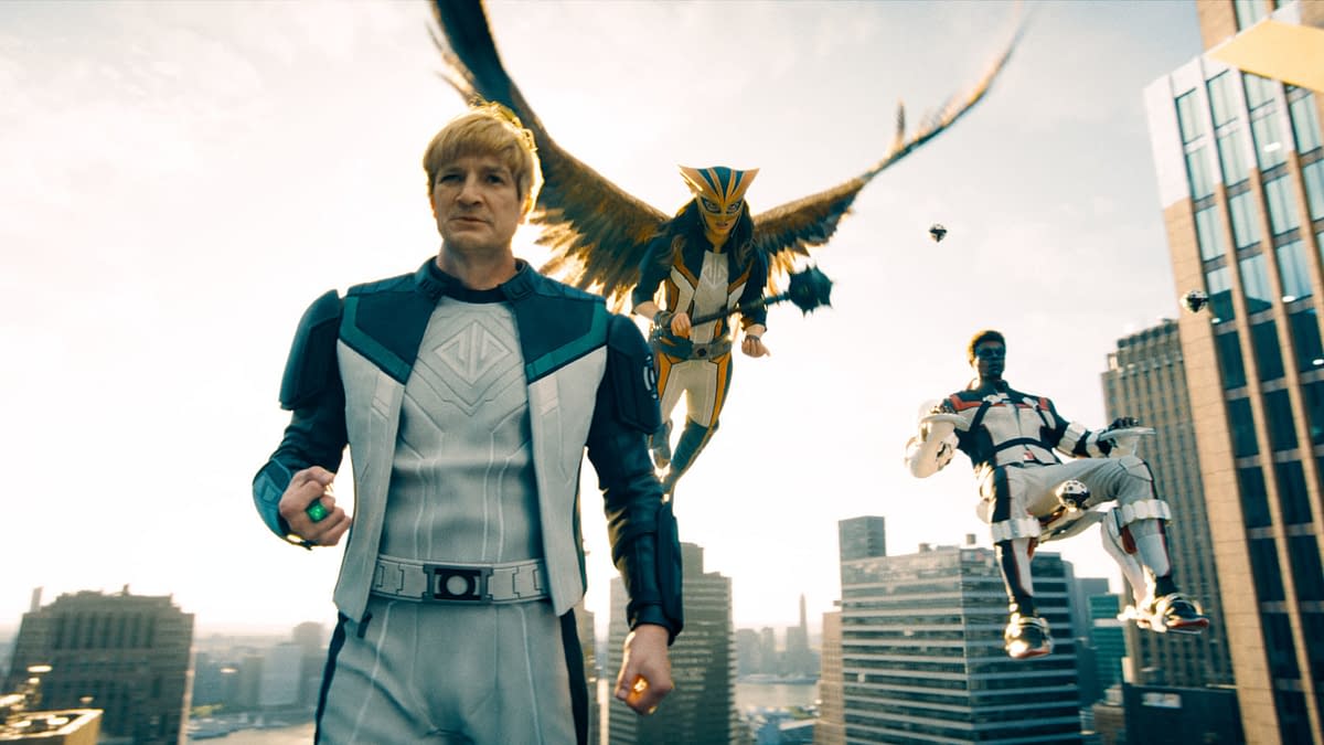 A scene from a superhero film featuring a main character in a futuristic outfit with a glowing green object, flanked by two flying characters, one with bird-like wings and the other seated in a high-tech chair, set against a city skyline.