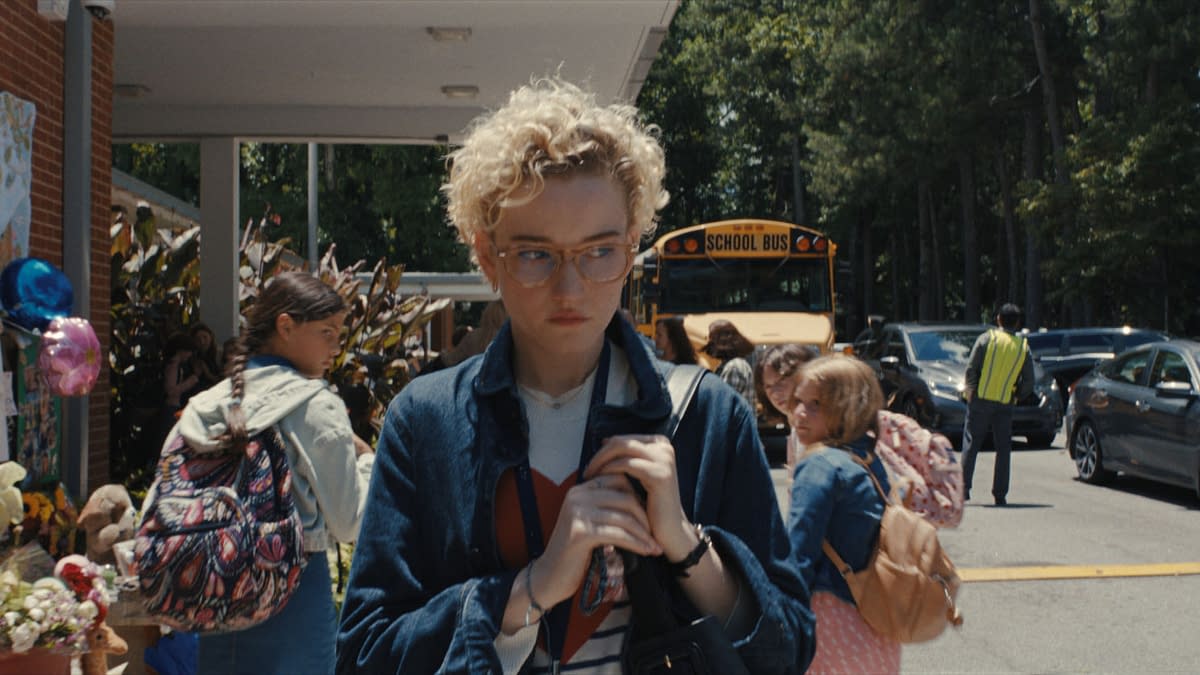 A scene outside a school showing a young woman with curly hair and glasses, looking somber while holding a bag. The background features students and a yellow school bus, with a memorial display reading "LOOK STRONG".