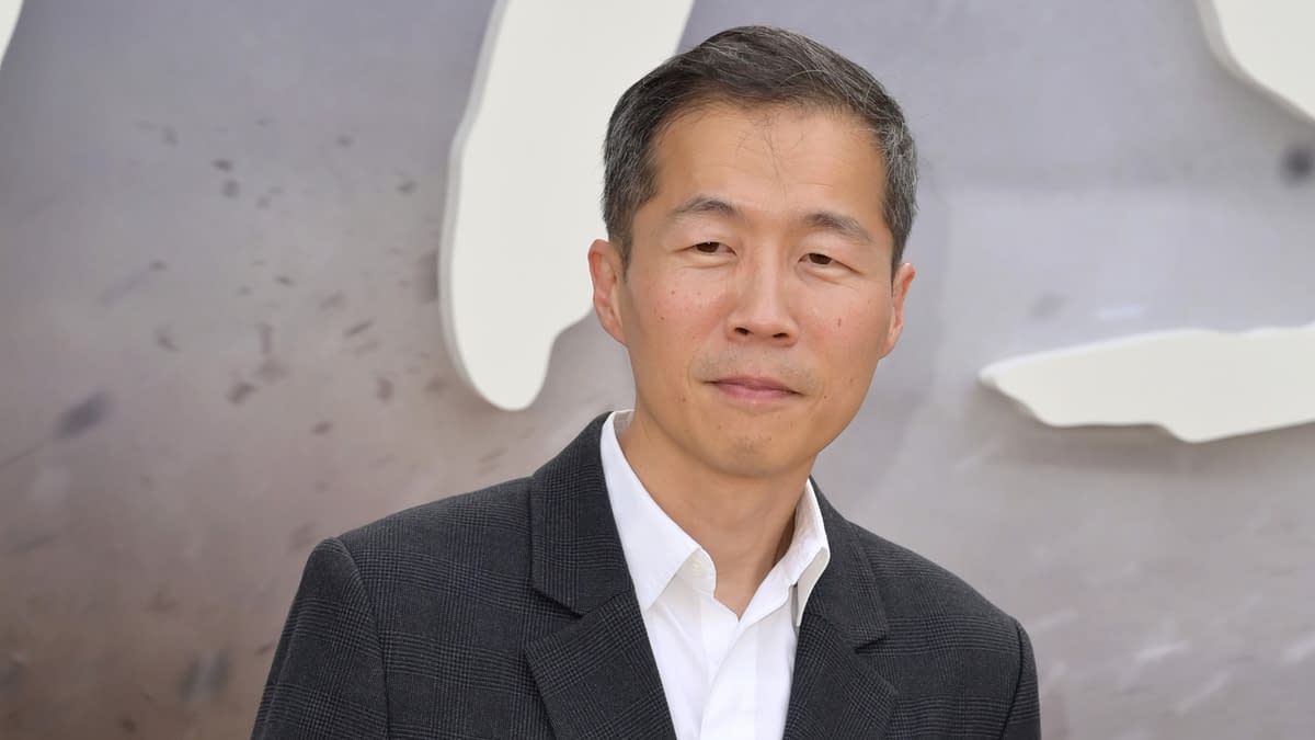 A man with short black hair is wearing a gray blazer over a white shirt, standing against a blurred background. He has a neutral expression and appears poised.