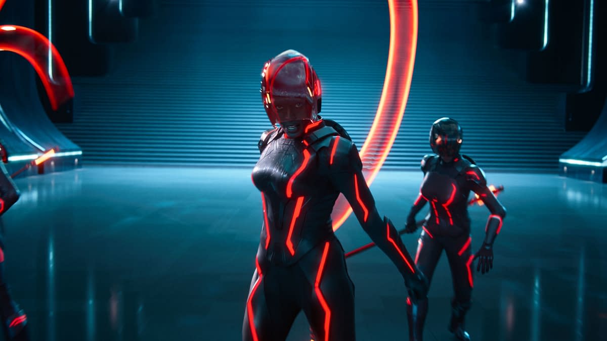 Jodie Turner-Smith as Athena stands confidently in a futuristic setting, wearing a sleek black suit with glowing red lines. Two other characters in similar outfits accompany her, set against a backdrop of illuminated structures and a staircase.