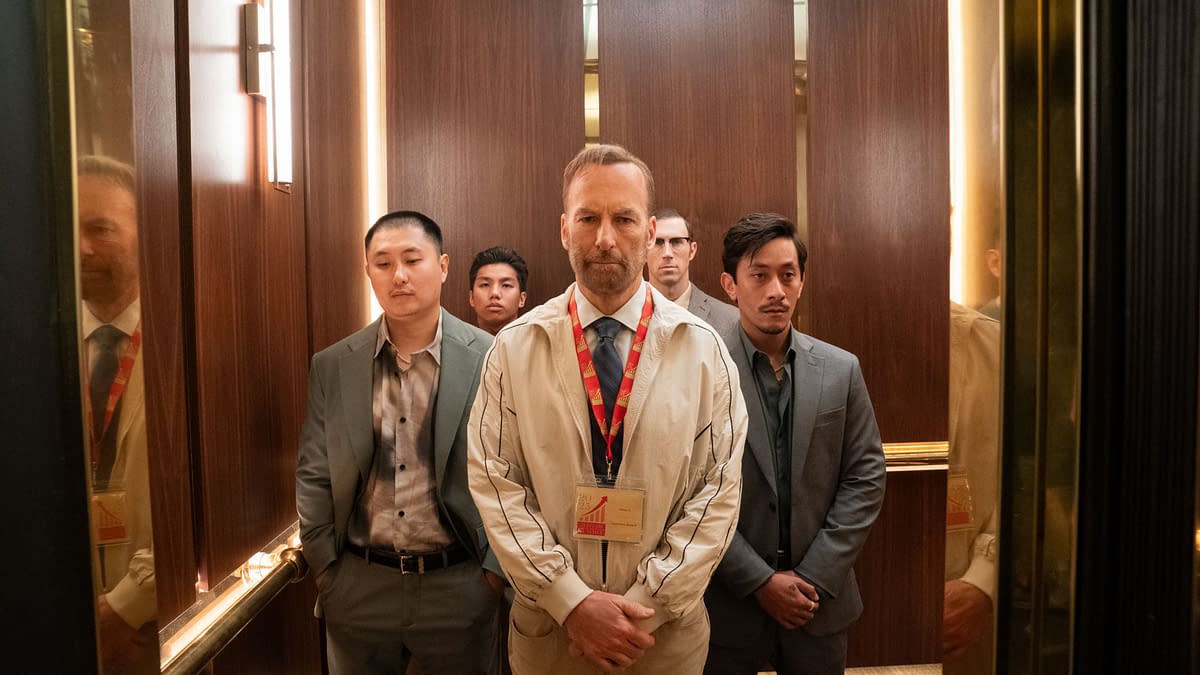 A group of five men stands solemnly in an elevator, dressed in suits and casual attire, with one man prominently wearing a badge around his neck. The setting features polished wooden walls and soft lighting, creating a tense atmosphere.