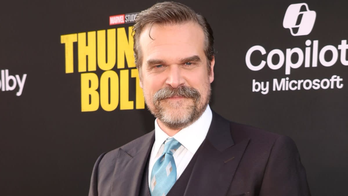 David Harbour smiles at the premiere of 'Thunderbolts,' dressed in a dark suit with a light blue tie, set against a black backdrop featuring the movie title.
