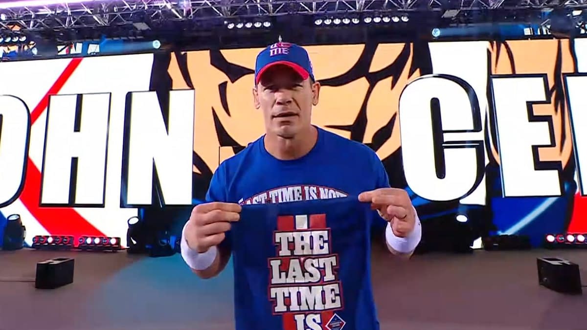 John Cena walks to the wrestling ring wearing a blue shirt that says "THE LAST TIME IS" while displaying a small item in his hand. The background features large graphics of his name.
