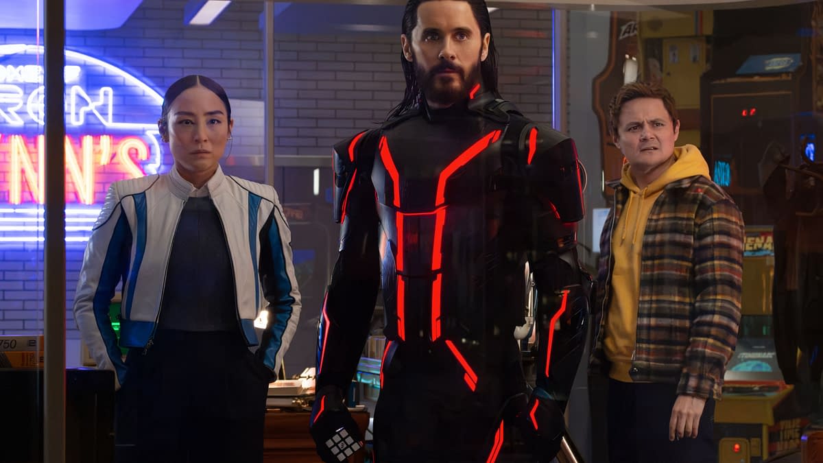 In a scene from Disney's live-action 'TRON: ARES', Greta Lee portrays Eve Kim, standing beside Jared Leto as Ares in a striking black suit with red accents, while Arturo Castro as Seth Flores looks on, all set against a vibrant arcade backdrop.