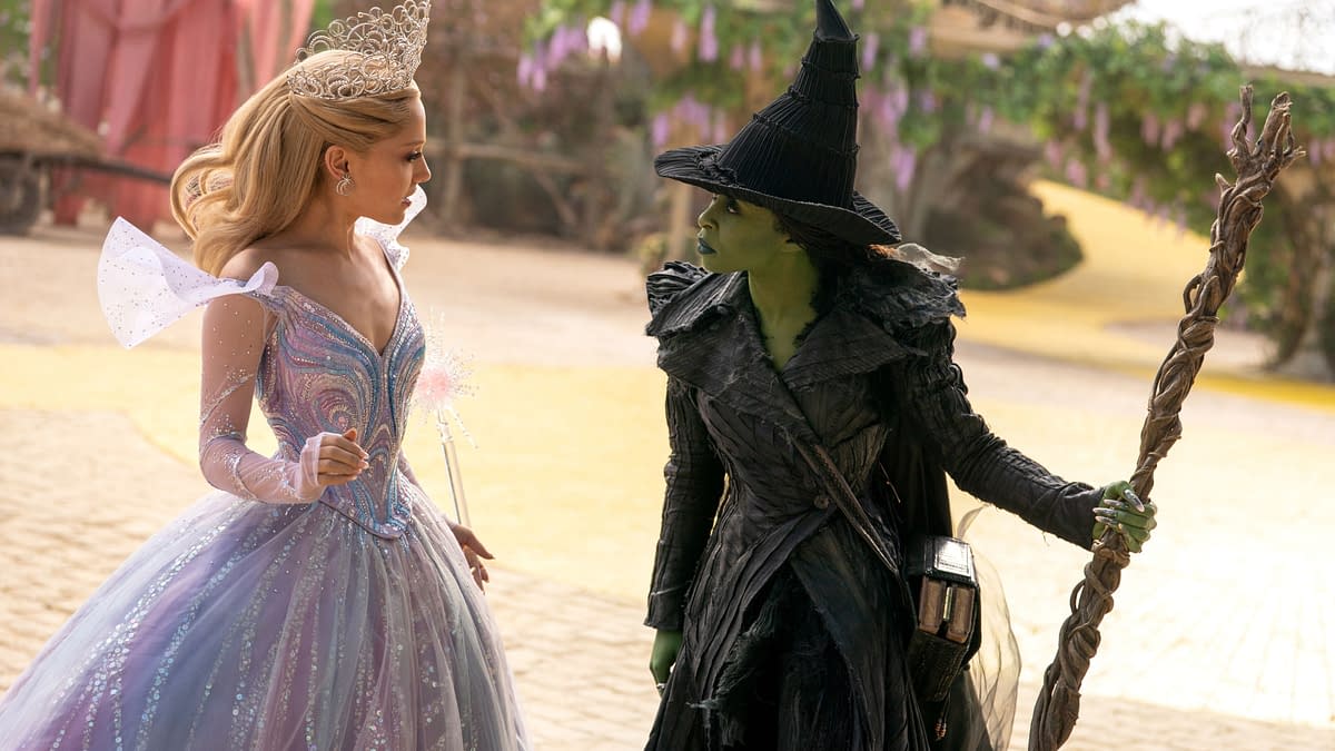 A promotional still from 'WICKED FOR GOOD' featuring Ariana Grande as Glinda in a sparkling gown and Cynthia Erivo as Elphaba, dressed in a black cloak and pointed hat. The scene captures a tense interaction between the two characters within a colorful, fantastical setting.