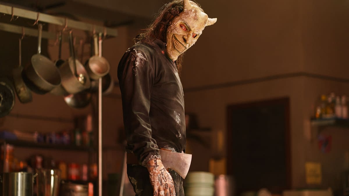Ethan Hawke portrays The Grabber in Black Phone 2, wearing a menacing mask and holding an axe, set in a dimly lit kitchen environment.