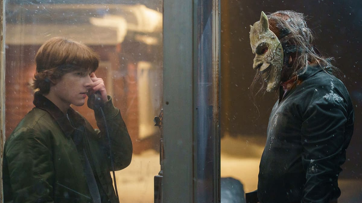 A tense confrontation occurs between a young man speaking on a payphone and a figure wearing a grotesque mask, set against a snowy backdrop, hinting at a suspenseful scene.