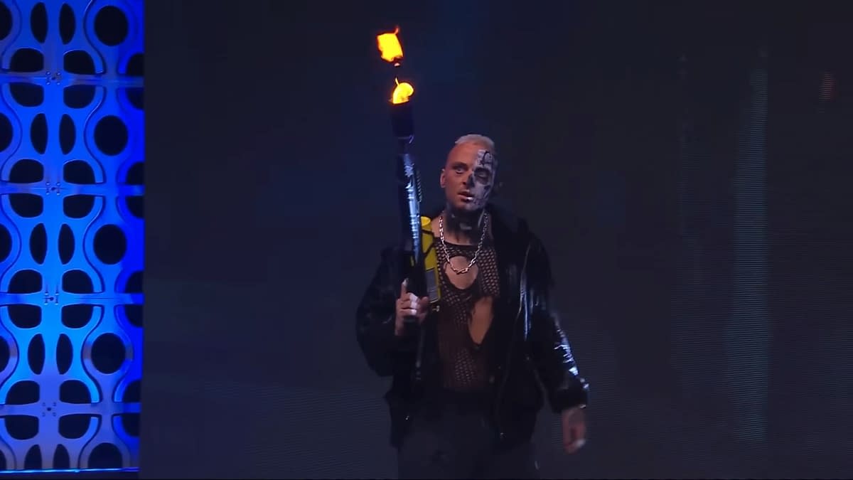 A wrestler with a heavily tattooed face and a dramatic outfit, holding a flamethrower that emits flames, appears on stage in low light, surrounded by a blue backdrop.