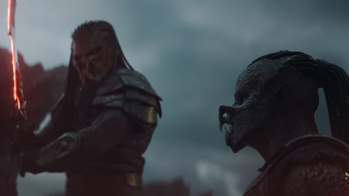 A scene from the film 'Predator: Badlands' features two alien characters, Kwei and Dek, dressed in armor. Kwei holds a glowing sword, while Dek observes amidst a dramatic cloudy background.