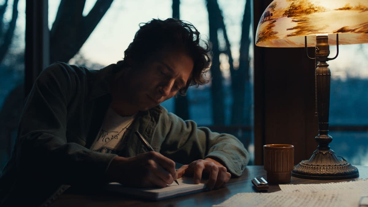 Illustration of Jeremy Allen White portraying Bruce Springsteen, seated at a table and writing in a notebook under a warmly lit lamp, with a window showing an outdoor setting in the background. This image is from 20th Century Studios' film 'SPRINGSTEEN: DELIVER ME FROM NOWHERE'.